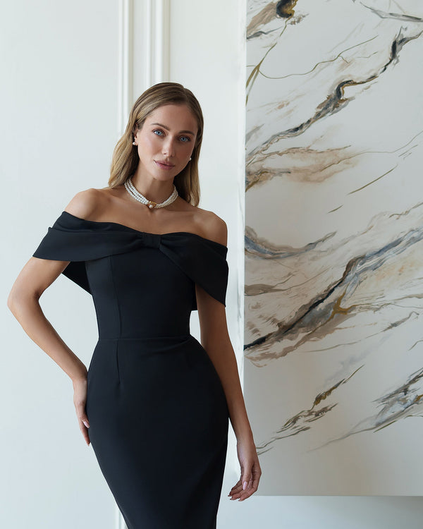 Woman in a black off-shoulder dress standing against a marble wall.