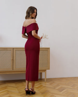 Woman in a burgundy off-shoulder dress standing in a room with wooden flooring and a light-colored wall.